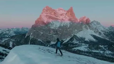 A jagged limestone peak in pink alpenglow; a lone figure walks the snow ridge below.
