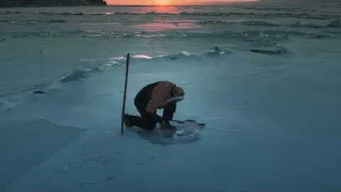 A figure in an orange jacket kneeling over a hole cut in cracked lake ice at sunset.